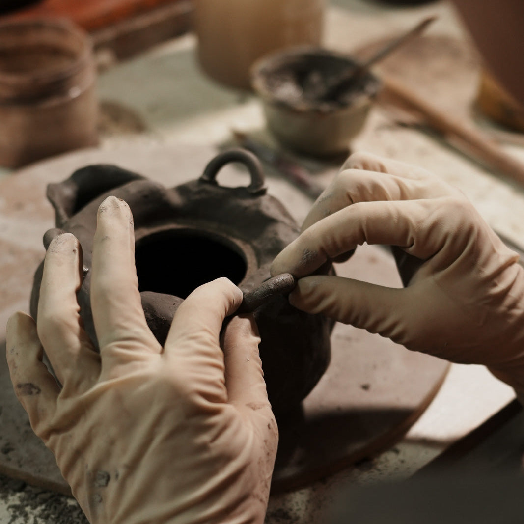Close-up of gloved hands shaping a dark clay vessel on a pottery workbench with tools and jars in the background.