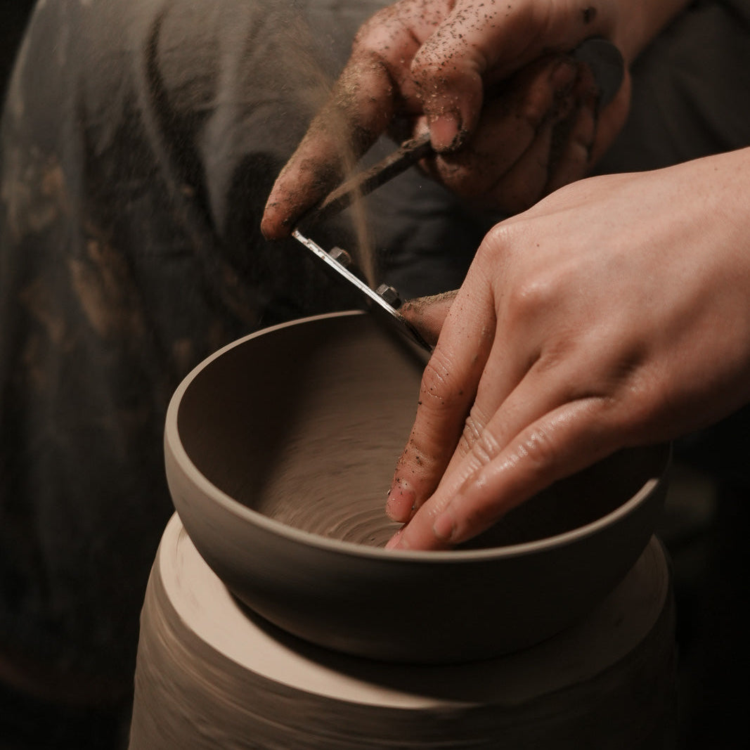 A close-up of a person's hands shaping a clay bowl on a pottery wheel, with the spinning clay wheel and the artisan's hands clearly visible.