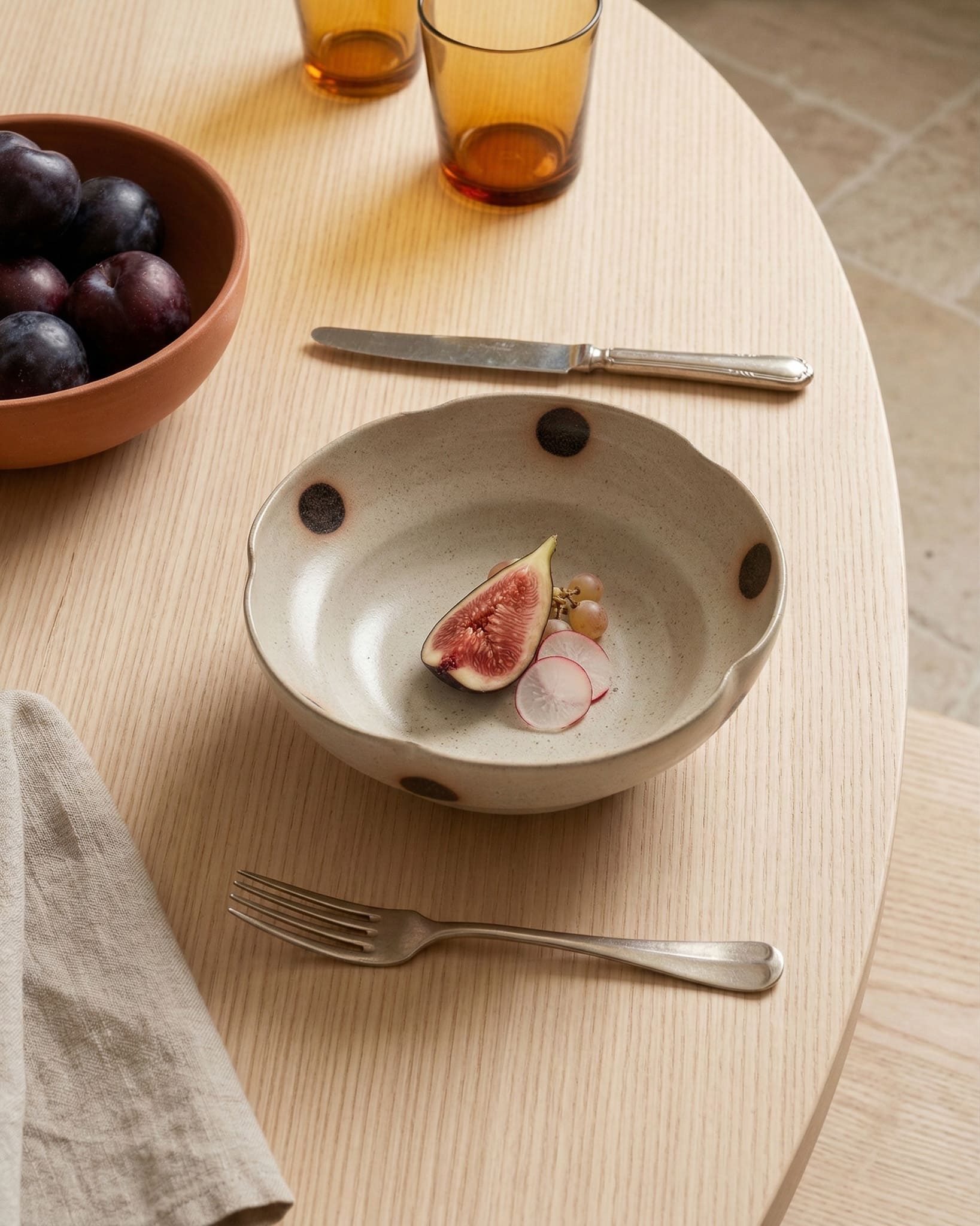 Table setting with a bowl containing figs, radishes, and grapes on a wooden table.