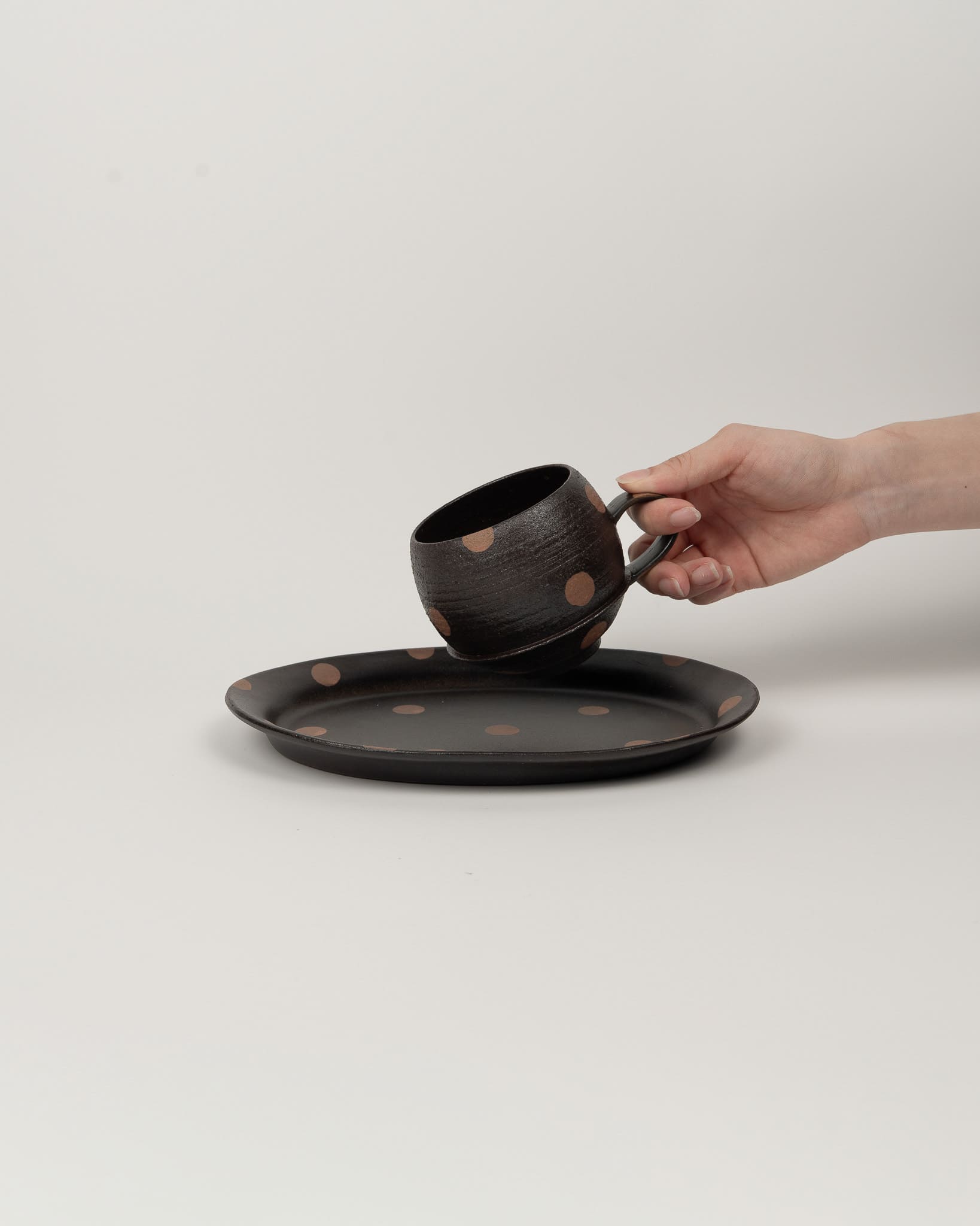 Hand holding a black ceramic cup with a matching saucer on a light gray background