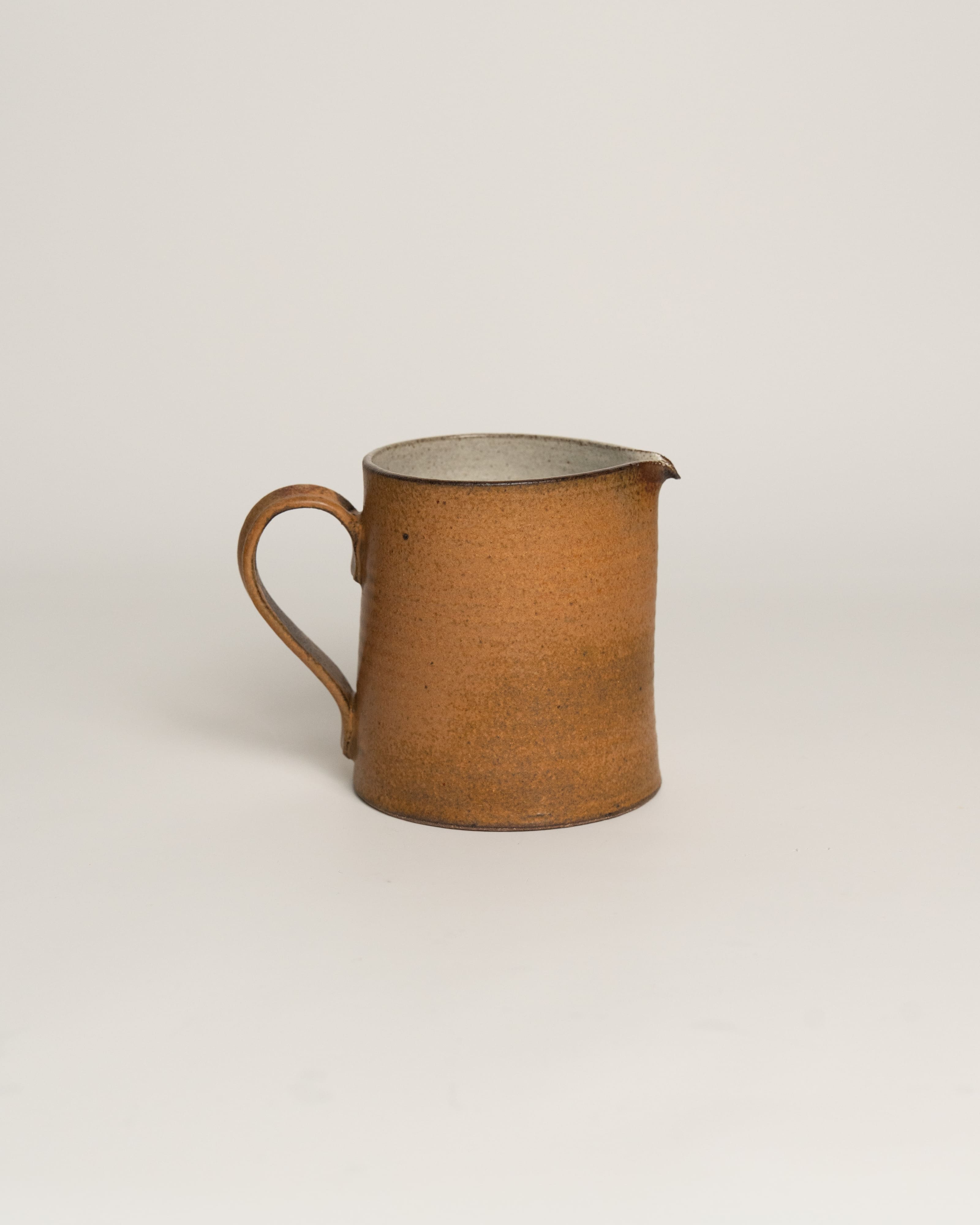 A rustic handmade ceramic pitcher with a warm brown speckled glaze, a curved handle, and a small pouring spout, set against a plain light gray background.