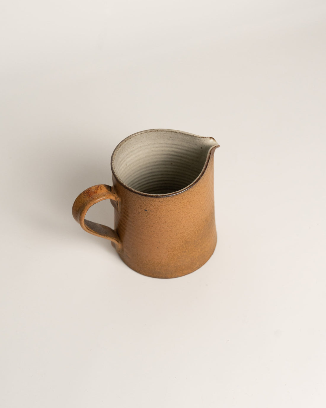 A rustic handmade ceramic pitcher with a warm brown speckled glaze, a curved handle, and a small pouring spout, set against a plain light gray background.