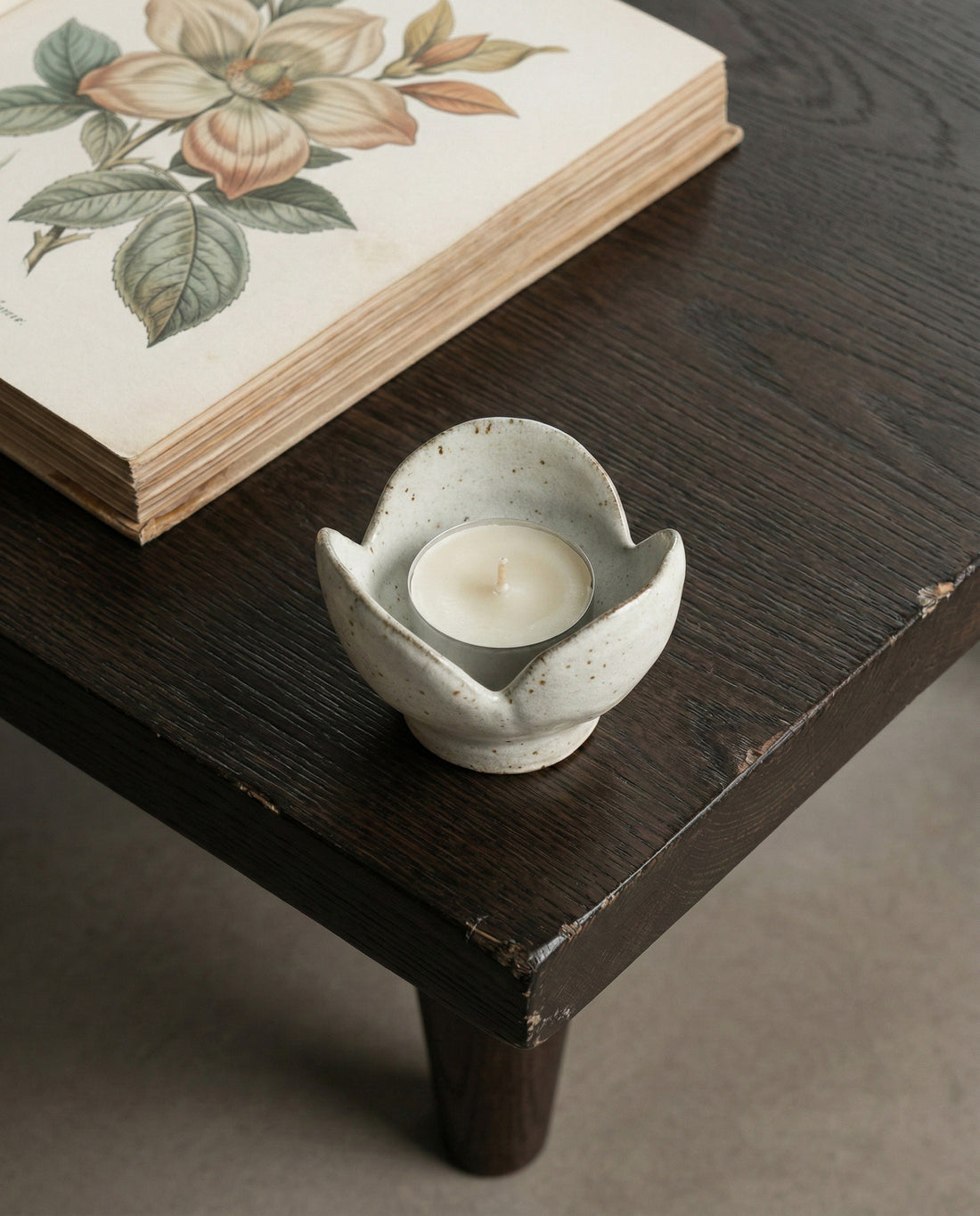 Ceramic candle holder with a candle on a wooden surface next to a book with floral design.