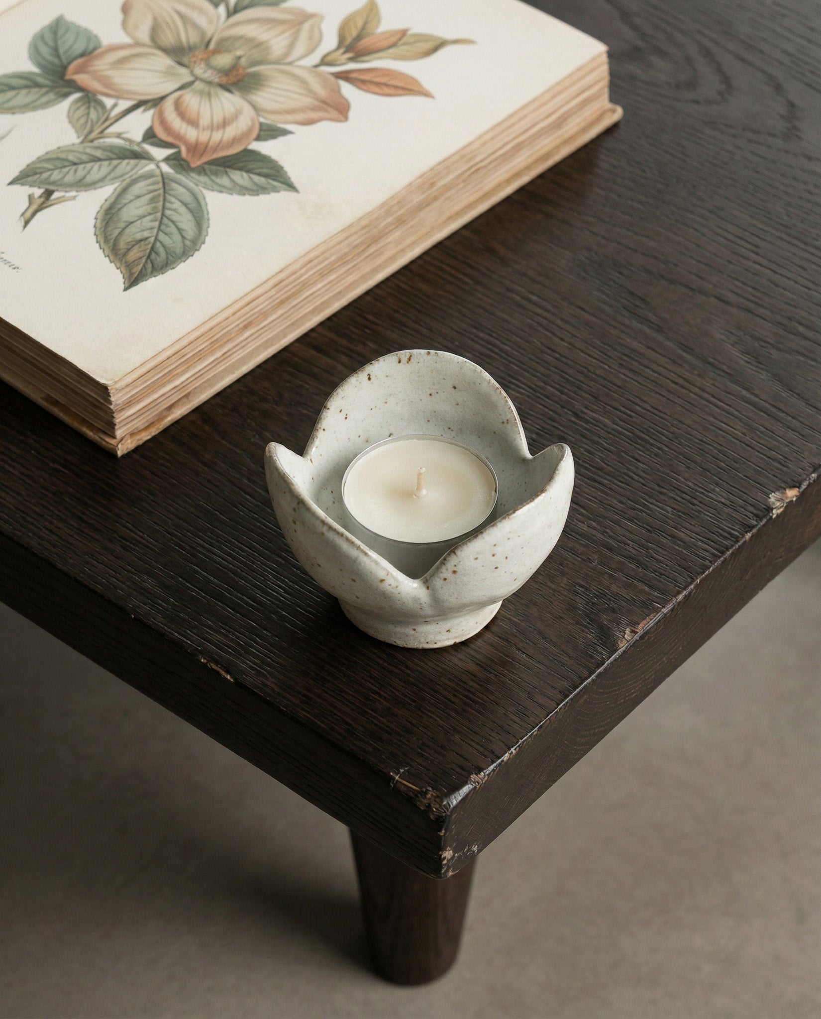 Ceramic candle holder with a candle on a wooden surface next to a book with floral design.