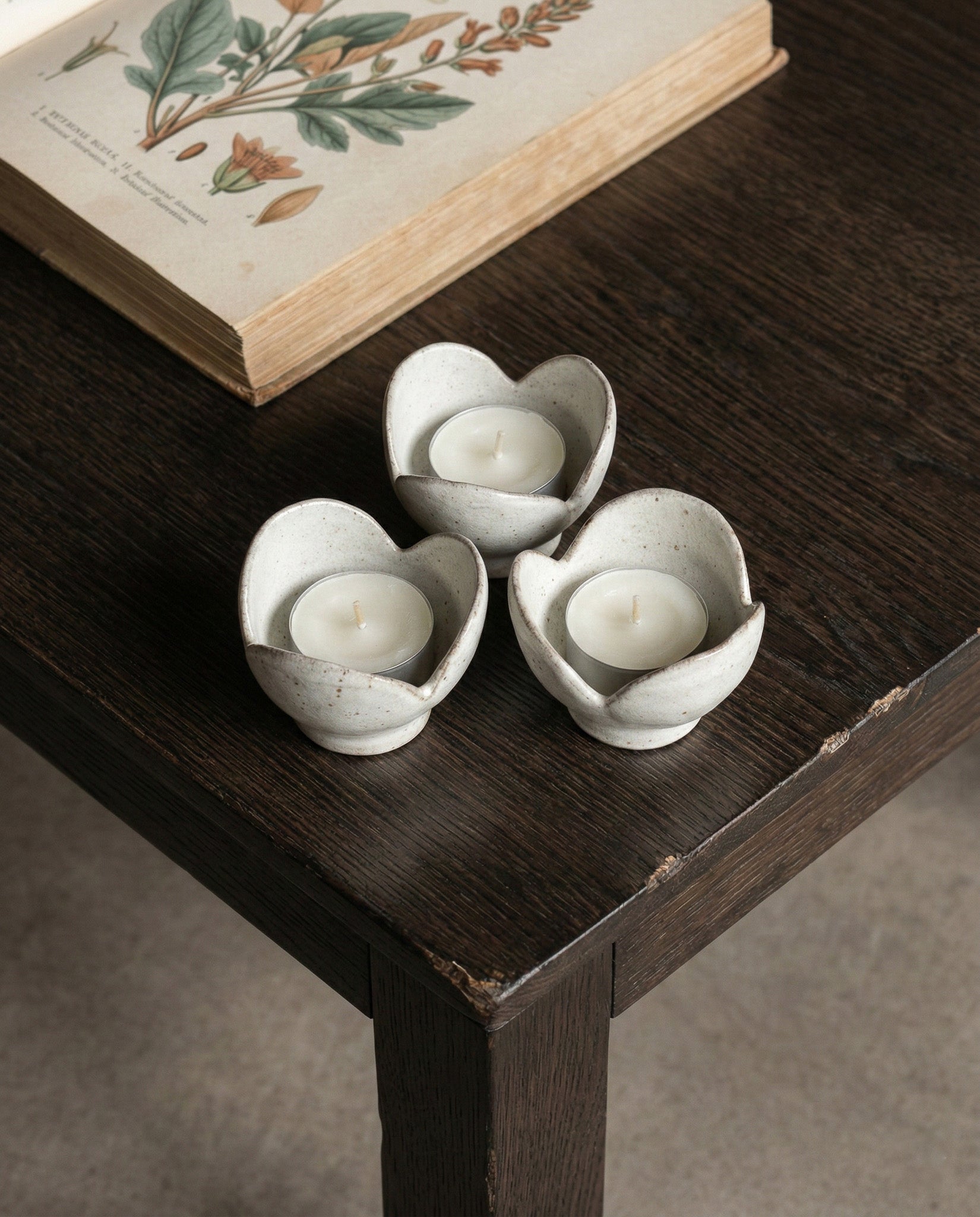Three ceramic flower-shaped candle holders with candles on a wooden surface, next to an open book with botanical illustrations.