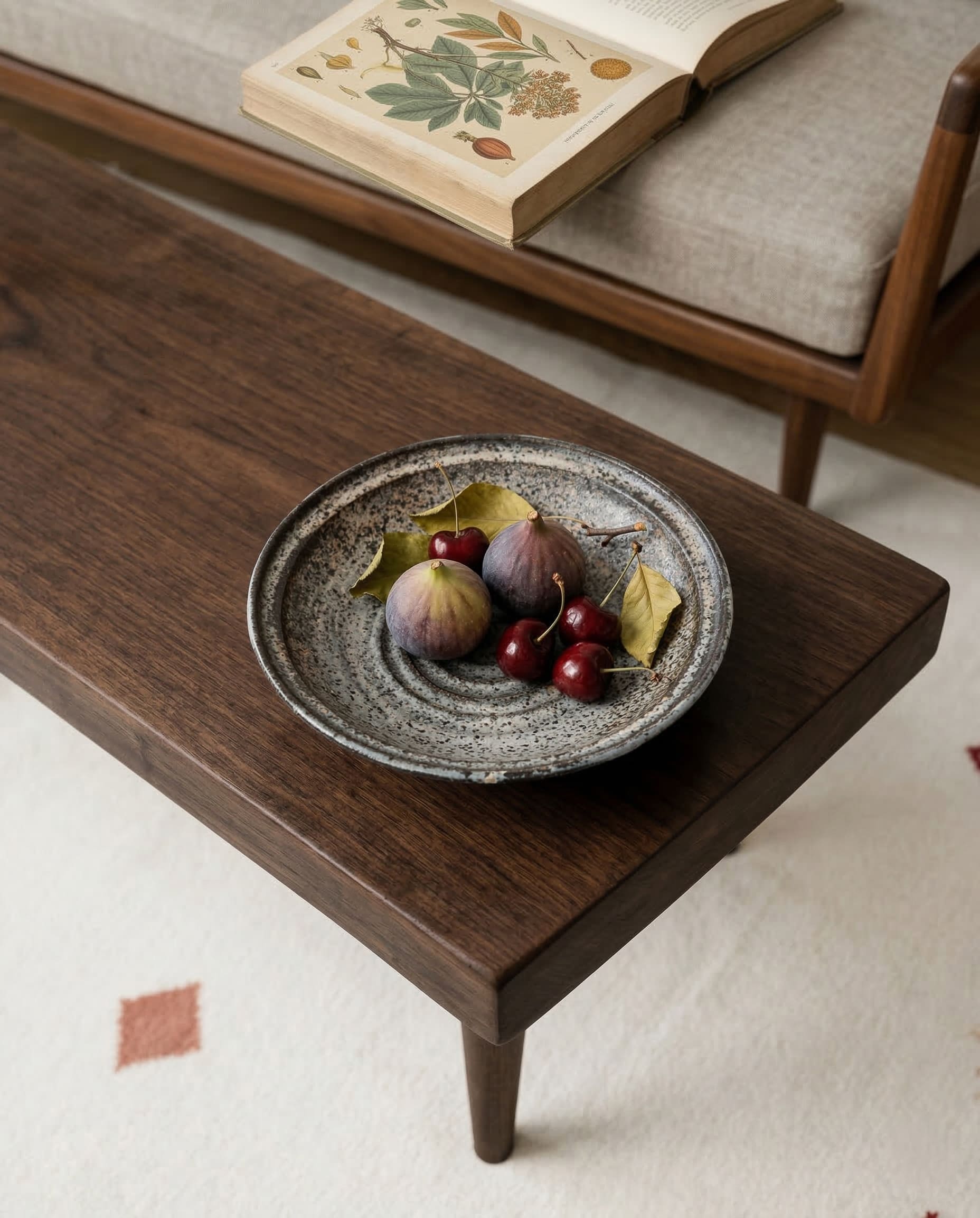 Wooden coffee table with a ceramic plate of figs and cherries, next to an open book on a sofa.