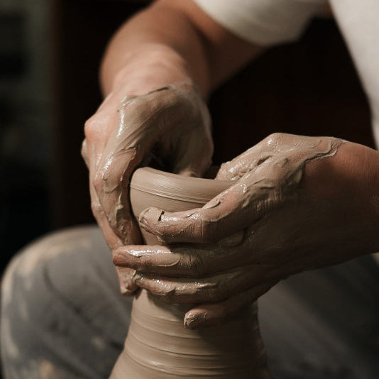 Close-up of a person's hands covered in clay shaping a pottery vase on a spinning wheel in a workshop.