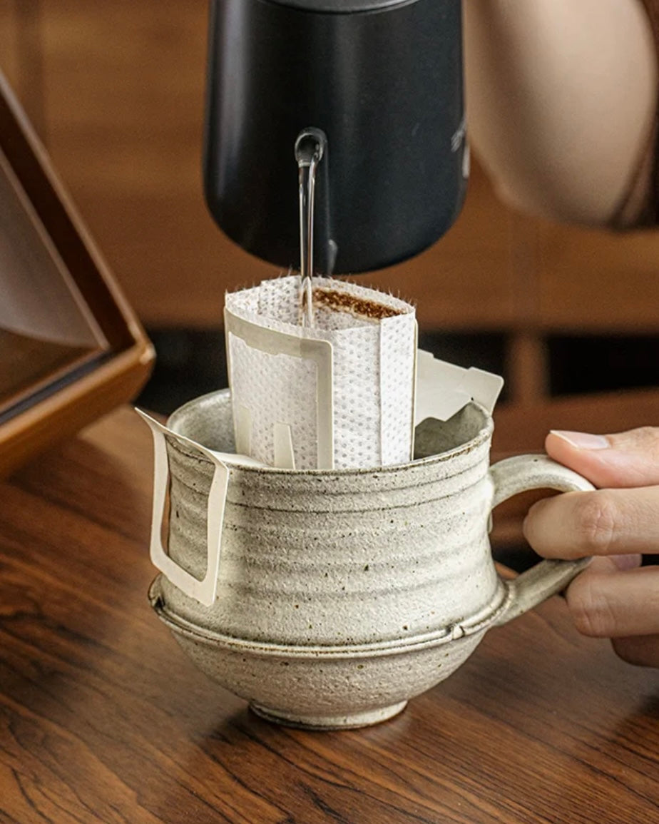 Person making coffee using a pour-over method with a ceramic mug and black coffee maker.