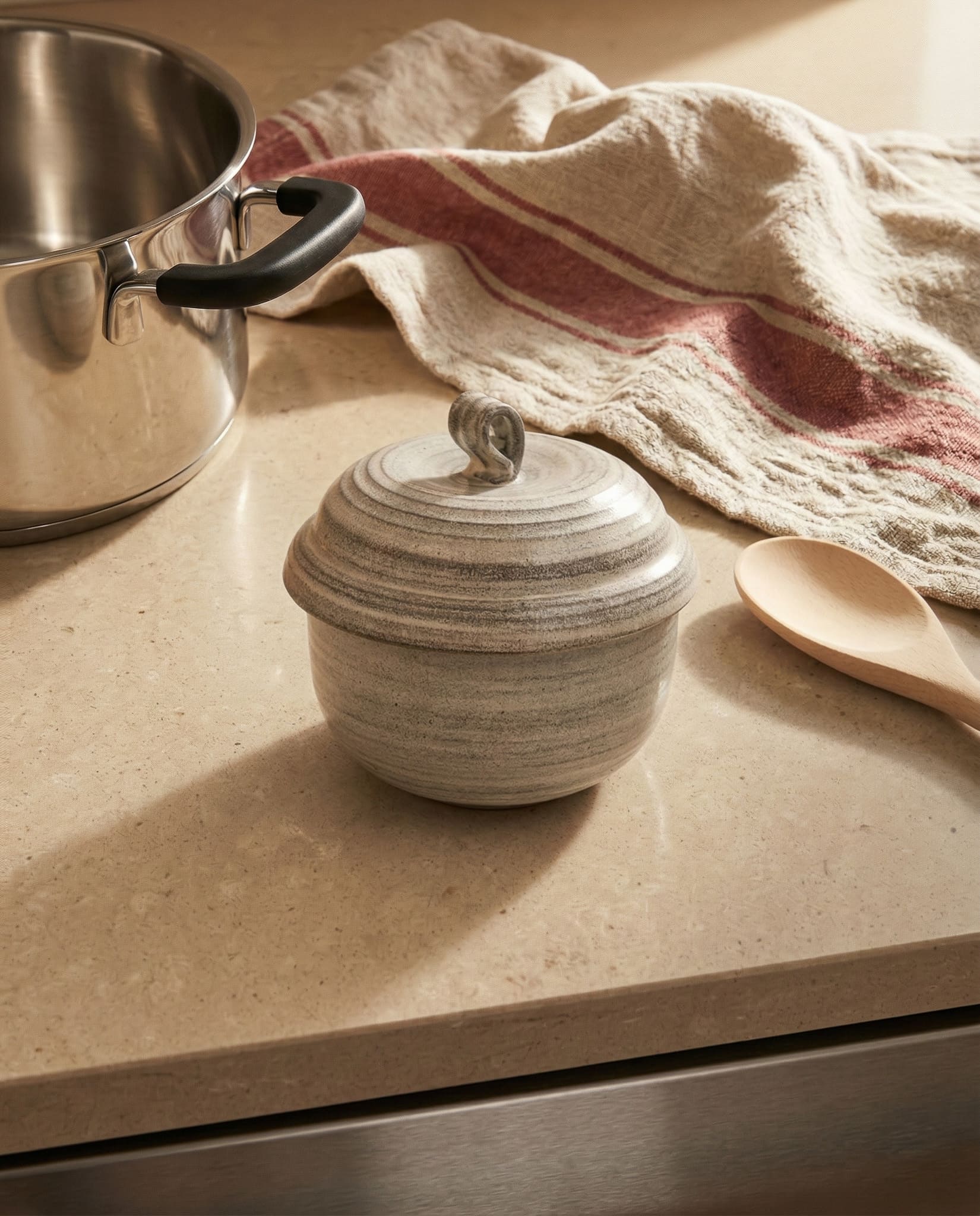 Small ceramic pot with a lid on a kitchen counter next to a towel and spoon