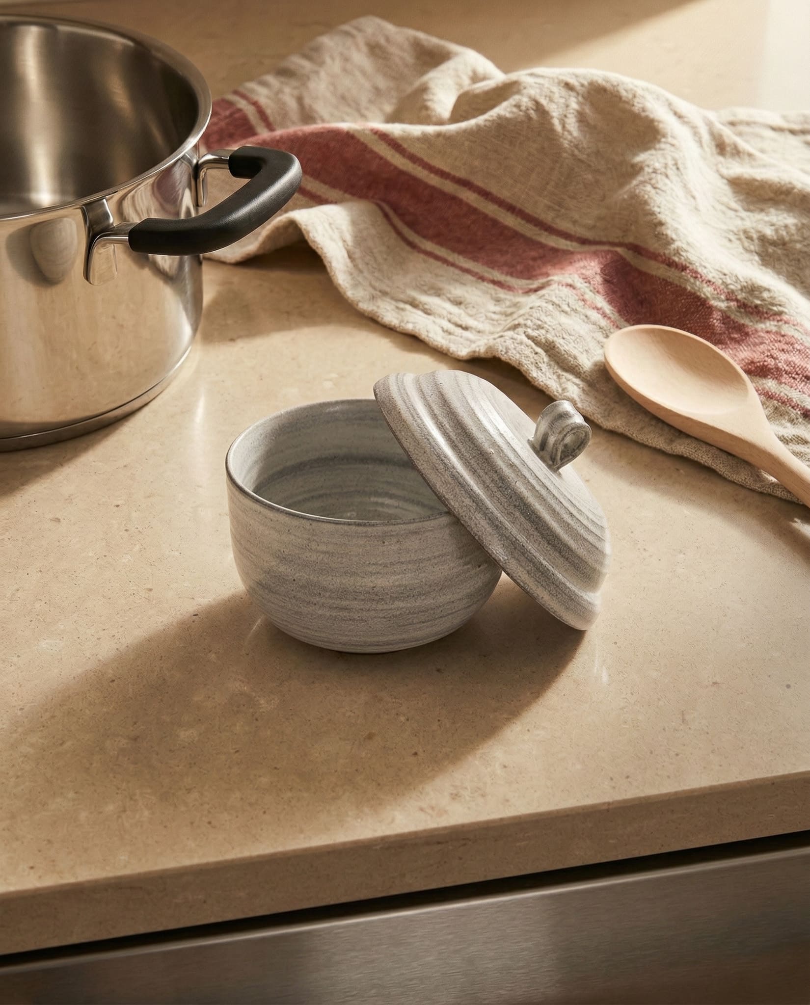 White ceramic bowl with a lid on a kitchen counter next to a pot and wooden spoon.