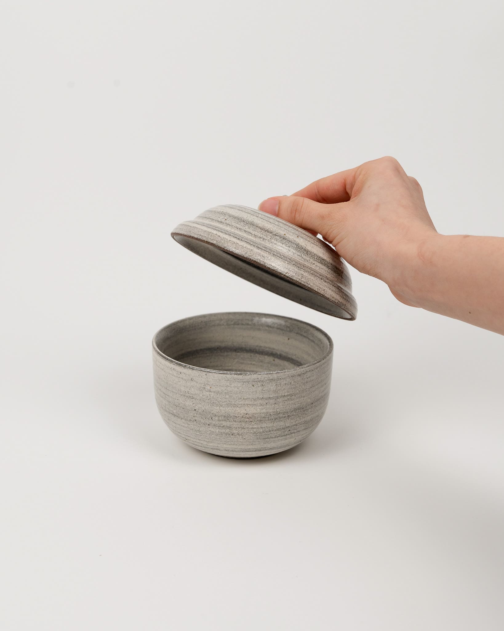 Hand holding a gray ceramic lid over a matching bowl on a white background