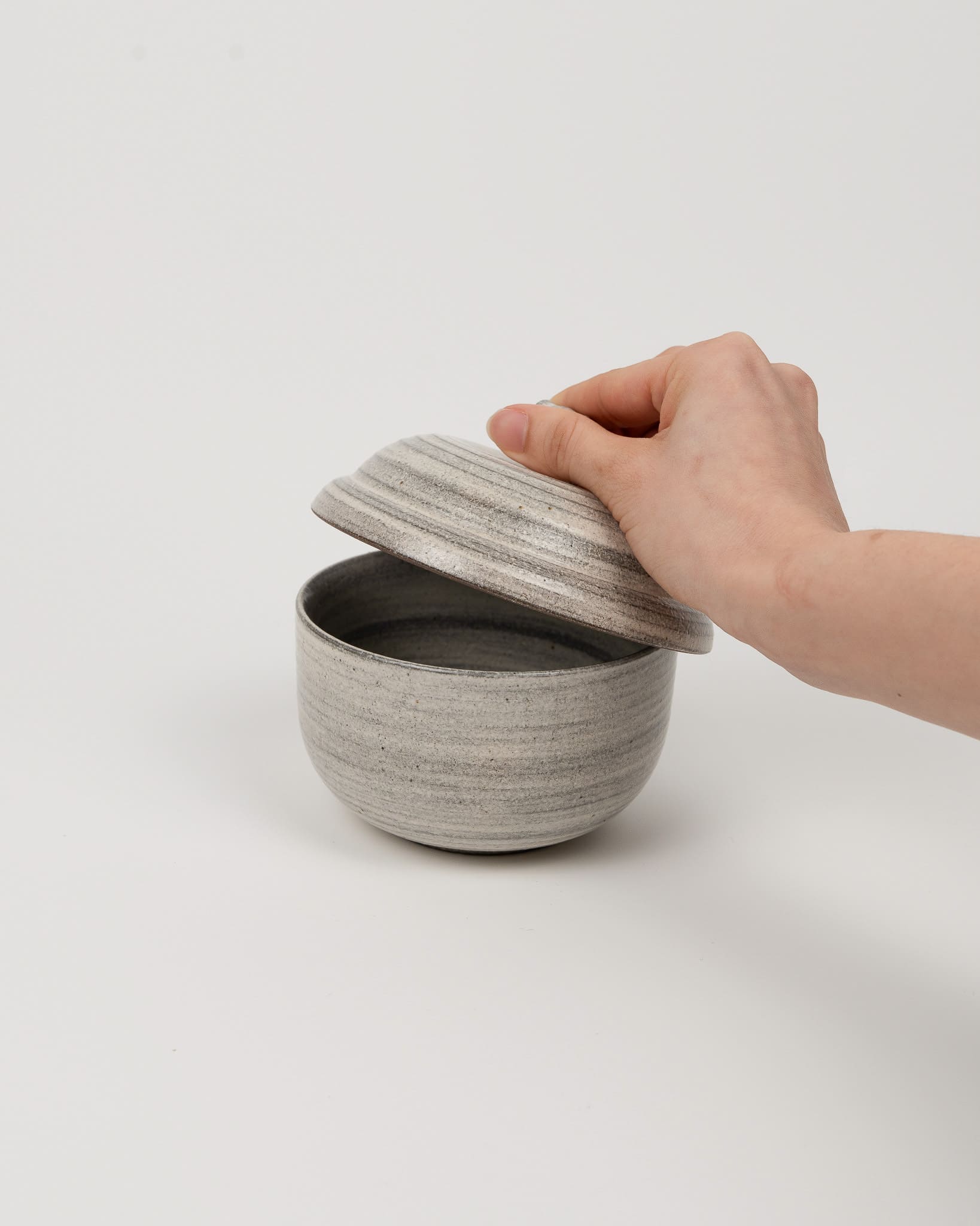 Hand holding a textured gray ceramic bowl with a matching lid on a light background