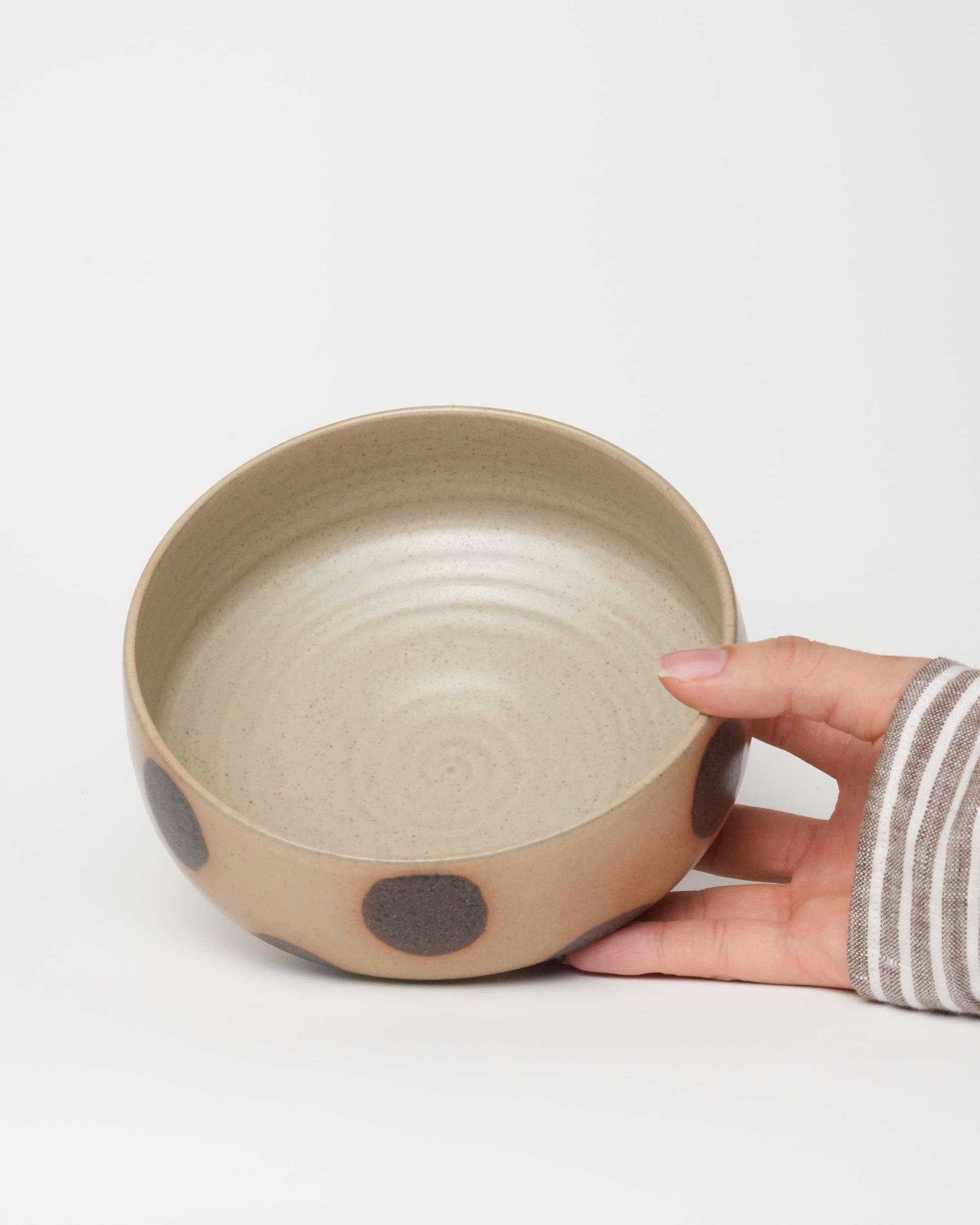 Hand holding a beige ceramic bowl with a brown dot on a light gray background