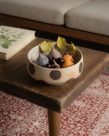 Bowl with fruits on a wooden table next to an open book, with a patterned rug underneath.
