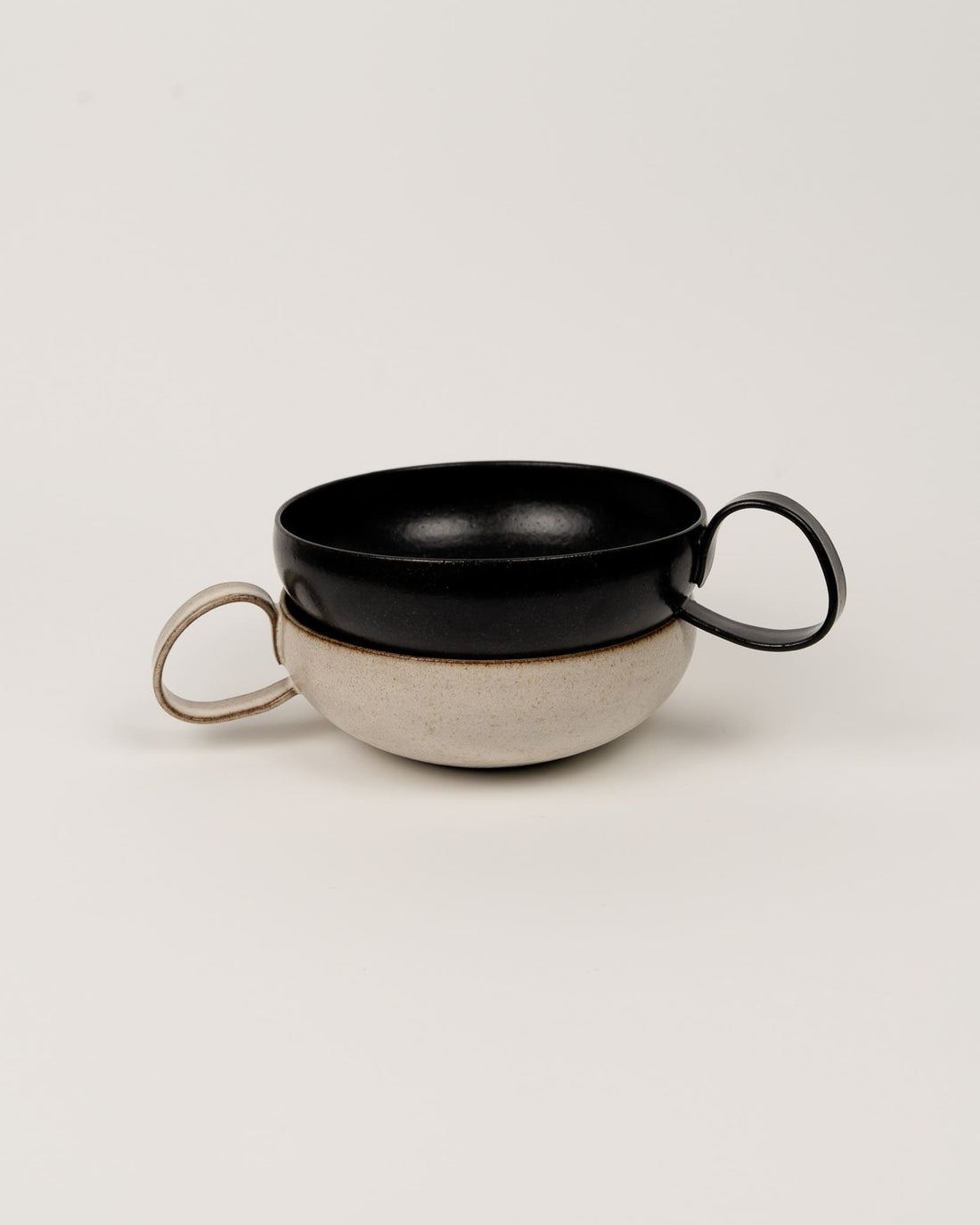 Two ceramic bowls, one black and one beige with handles, on a light gray background.