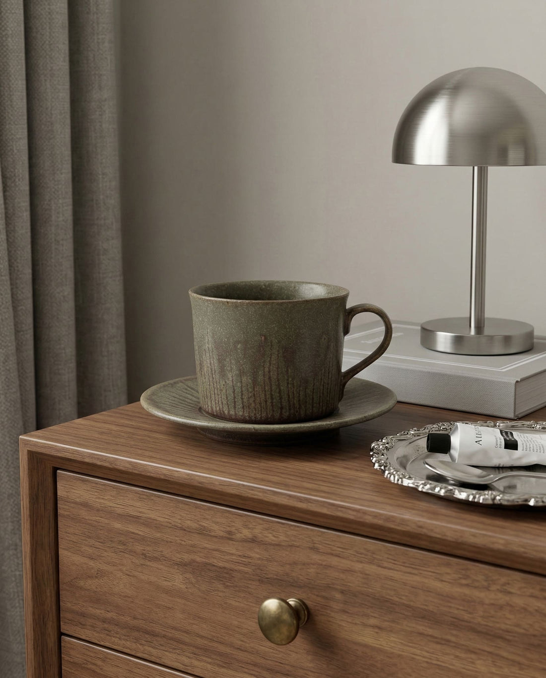 Ceramic cup and saucer on a wooden dresser with a lamp and decorative items.